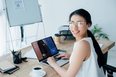 neşeli latin iş kadını kamera gülümsüyor iken ekranda rezervasyon web sitesi ile dizüstü bilgisayar kullanarak