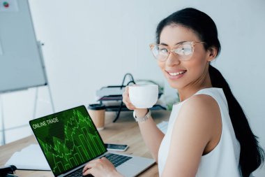 kahve fincanı tutarken ekranda online ticaret web sitesi ile dizüstü bilgisayar kullanarak gülümseyen latin iş kadını