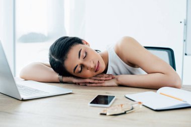 exhausted latin businesswoman sleeping at workplace near notebook and smartphone with blank screen clipart