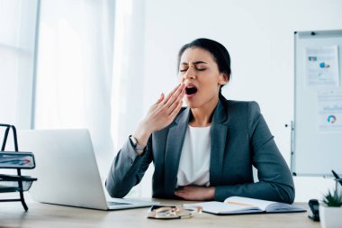 sleepy latin businesswoman yawning with closed eyes while sitting at workplace in office clipart