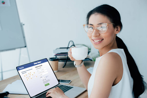 КИЕВ, УКРАИНА - 26 апреля 2019 года: Улыбающаяся латинская бизнесвумен с помощью ноутбука с сайта Youtube на экране, держа в руках чашку кофе
.