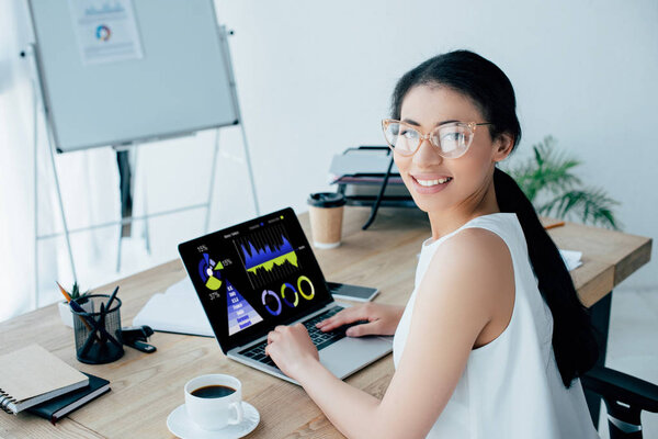 young latin businesswoman looking at camera while using laptop with graphs and charts on screen