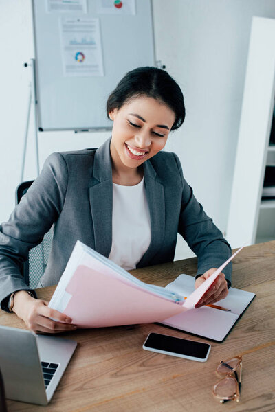 smiling latin businesswoman holding paper folder while sitting at workplace near smartphone with blank screen