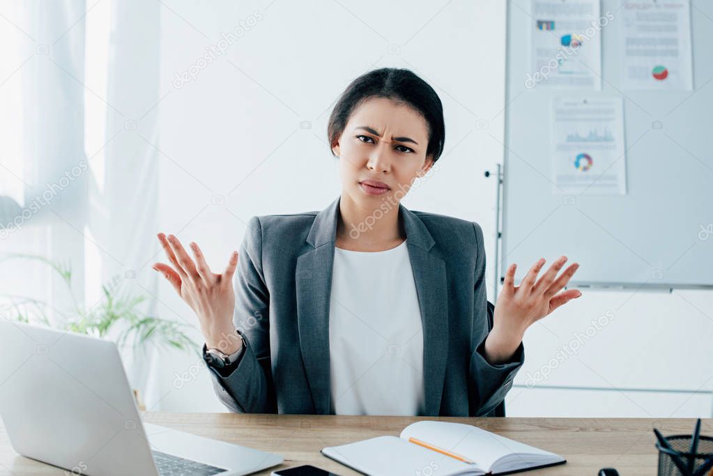 displeased latin businesswoman gesturing while sitting at workplace and looking at camera