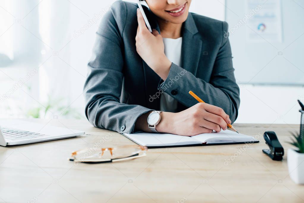 cropped shot of smiling latin businesswoman writing in notebook while talking on smartphone