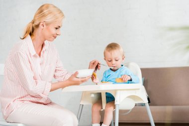 mutlu sarışın anne kase tutan seçici odak ve evde sevimli bebek oğlu besleme 