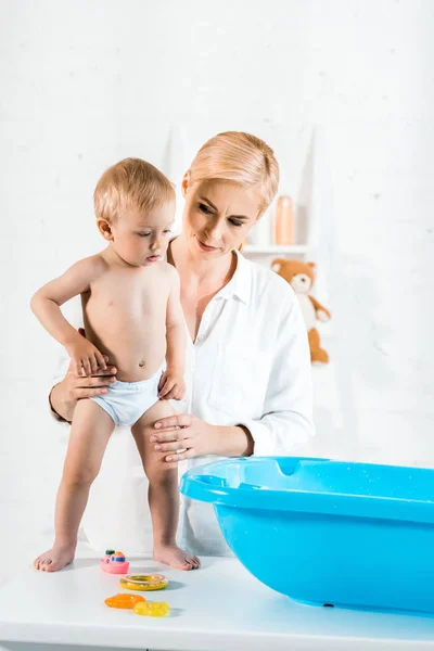 美丽的母亲看着婴儿浴缸 而抱着蹒跚学步的儿子在浴室 — 图库照片
