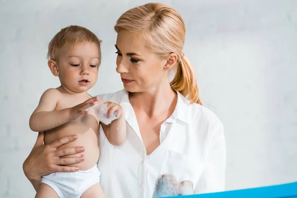 Attraktive Frau Hält Niedlichen Kleinkindsohn Den Armen Der Badeschaum Betrachtet — Stockfoto