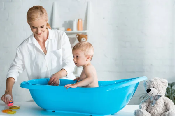开朗的母亲看着可爱的幼儿儿子在家里洗澡 — 图库照片