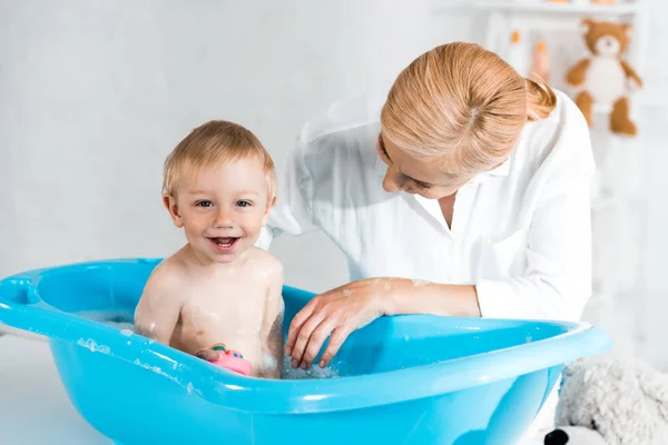 金发妈妈看着可爱的幼儿儿子微笑在蓝色婴儿浴缸 — 图库照片