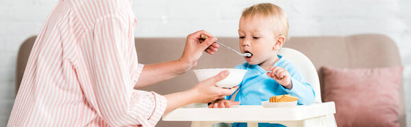 panoramic shot of mother holding bowl and feeding cute toddler son at home 
