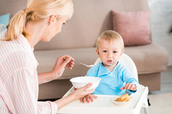 selective focus of blonde mother holding bowl and feeding cute toddler son at home 