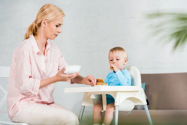 选择性的焦点金发母亲拿着碗附近可爱的幼儿儿子在喂食椅 — 图库照片