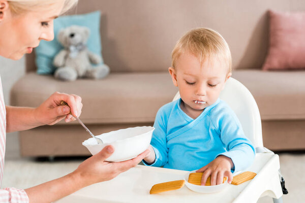 selective focus of mother holding bowl with baby food near cute toddler son at home 