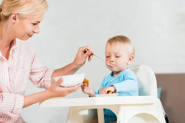 金发母亲拿着碗和喂养可爱的幼儿儿子在家里 — 图库照片