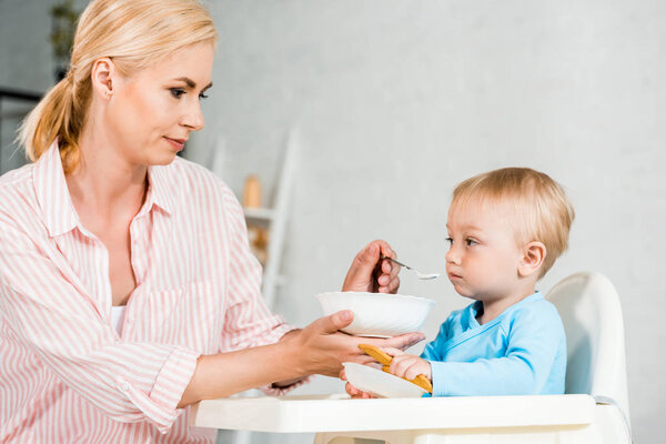 blonde mother holding bowl and feeding cute toddler kid at home 