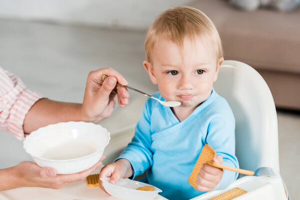 cropped view of mother holding bowl and feeding cute toddler son at home 