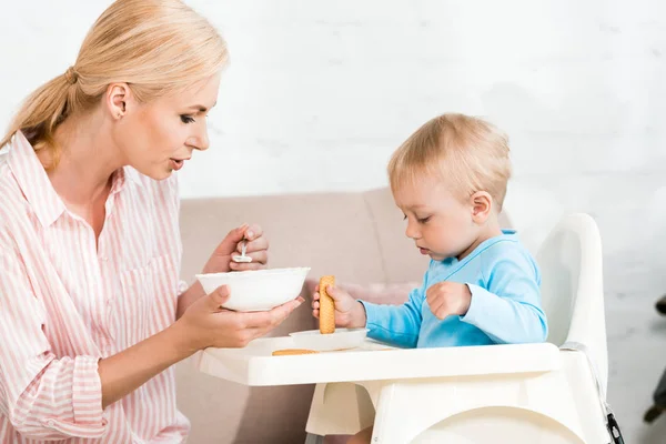 有吸引力的母亲拿着勺子与婴儿食品接近可爱的幼儿儿子坐在喂食椅 — 图库照片