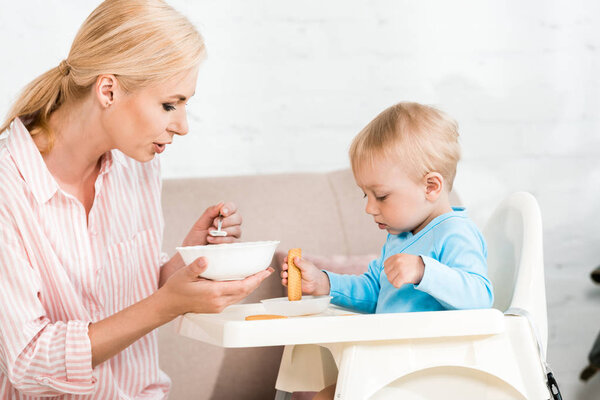 attractive mother holding spoon with baby food near cute toddler son sitting in feeding chair 