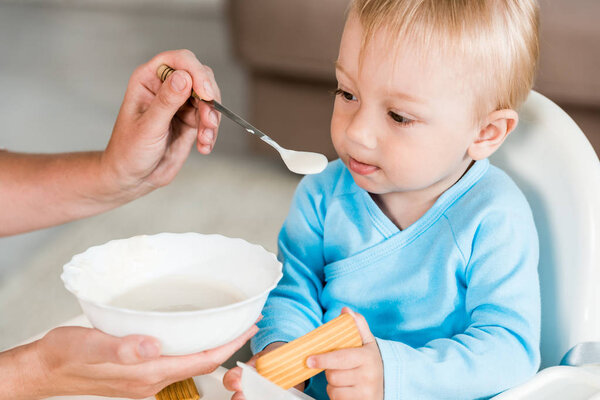 cropped view of mother holding spoon with baby food near cute toddler son sitting in feeding chair 