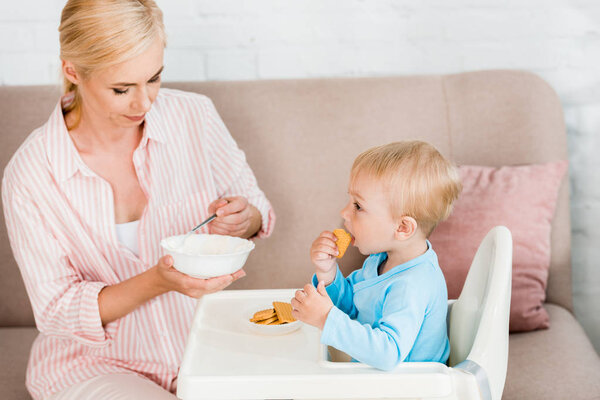 blonde mother holding spoon with baby food near cute toddler son sitting in feeding chair 