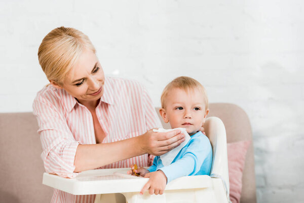 happy blonde mother holding napkin near cute toddler son sitting in feeding chair 