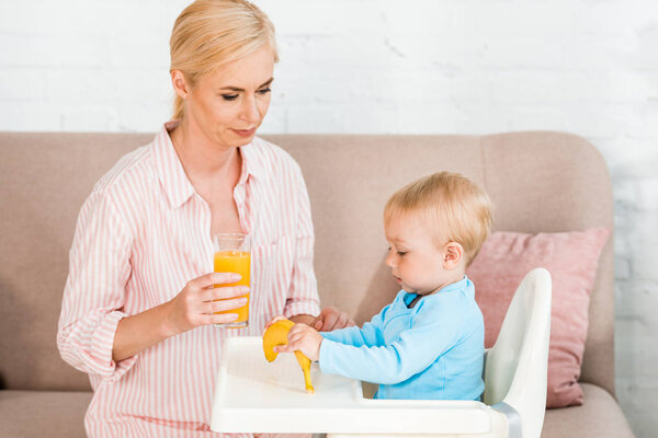 blonde mother holding glass with orange juice near cute toddler son sitting in feeding chair 