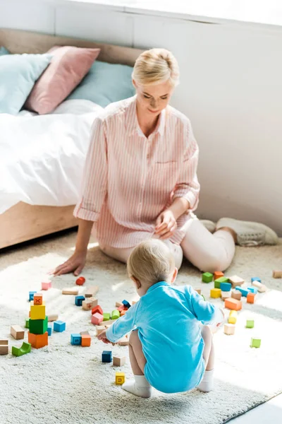 快乐的母亲坐在地毯上 看着蹒跚学步的儿子玩建筑砖 — 图库照片