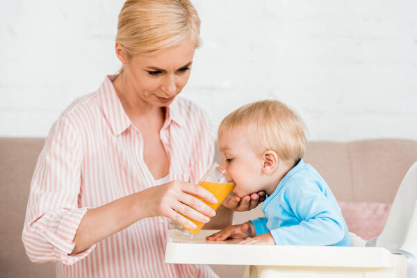 attractive blonde mother holding glass while toddler son drinking orange juice 