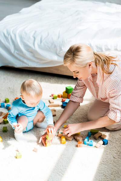 attractive blonde woman sitting on carpet near toddler son playing with building bricks 