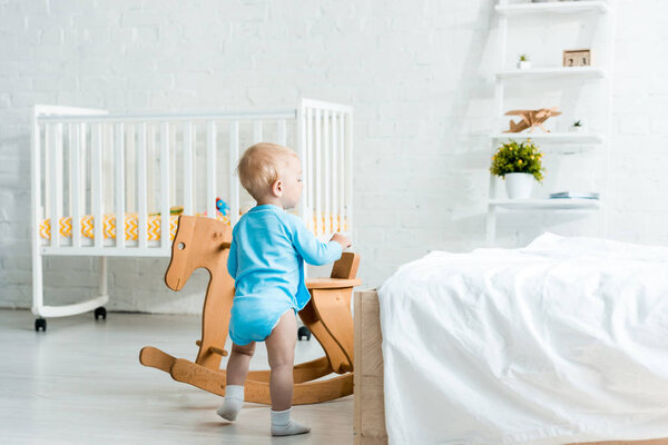 cute toddler child standing near wooden rocking horse in modern bedroom 