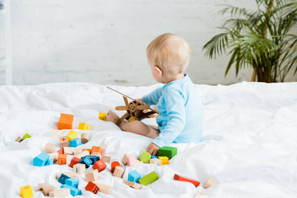 toddler boy playing with wooden biplane near colorful toy blocks on bed 