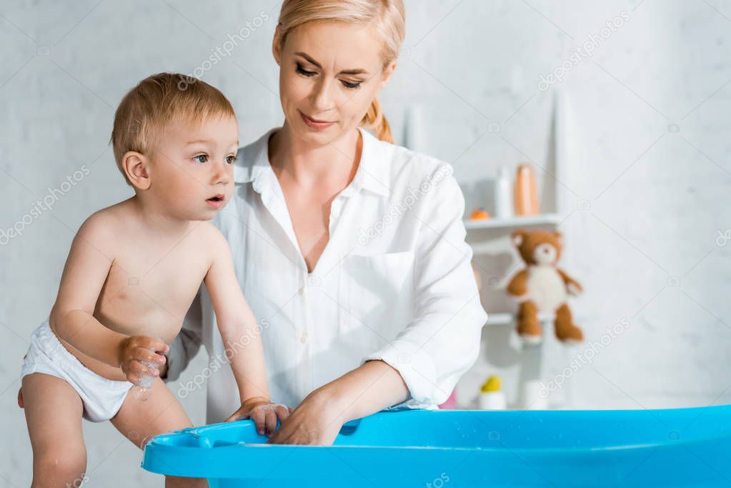 blonde woman standing and looking at blue baby bathtub near toddler son 