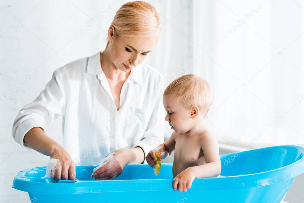 blonde mother standing near toddler kid in baby bathtub 