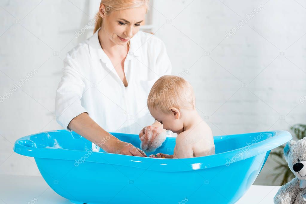 cheerful blonde mother smiling near cute toddler son taking bath 