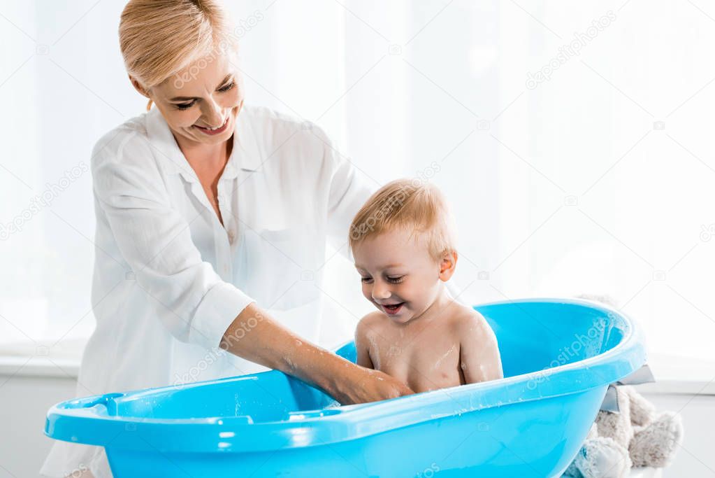 cheerful blonde mother smiling while washing cute toddler son in blue baby bathtub 
