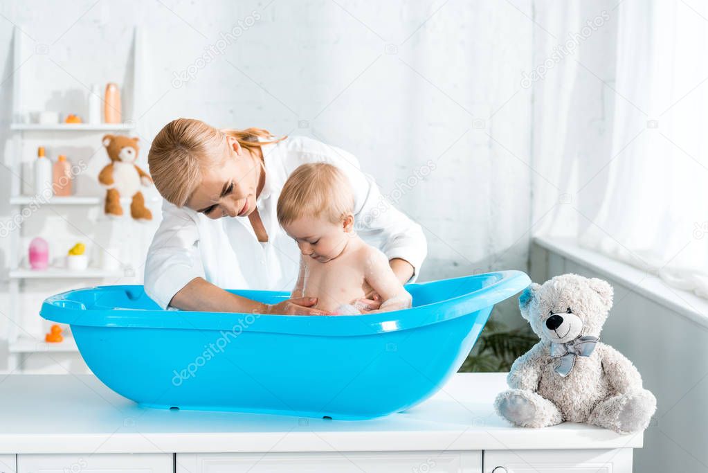 blonde cheerful mother washing happy toddler son in baby bathtub 