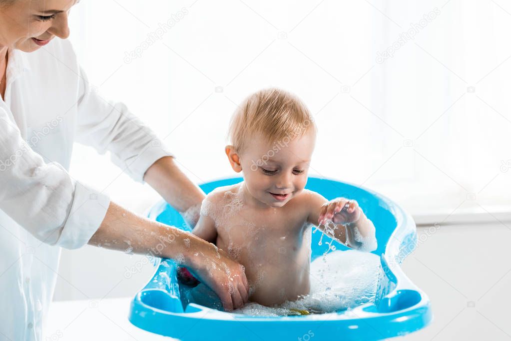 cropped view of happy mother washing toddler child in blue baby bathtub 