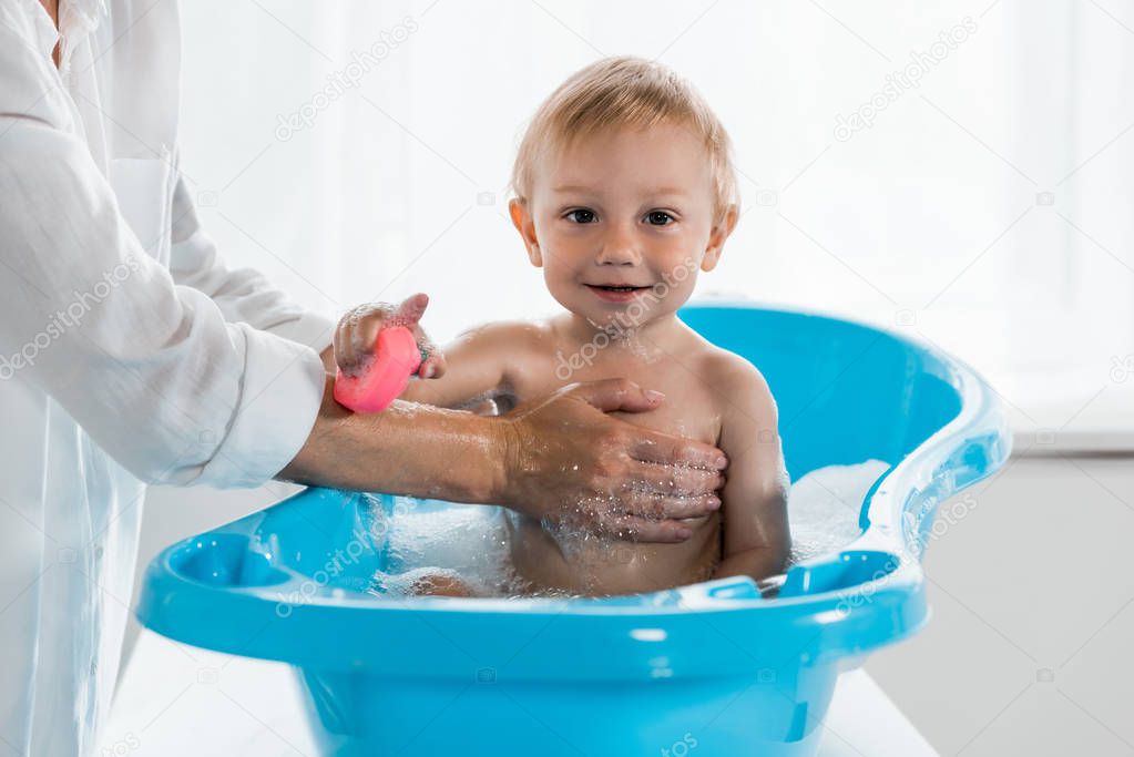 cropped view of mother washing happy toddler child with rubber toy in blue baby bathtub 