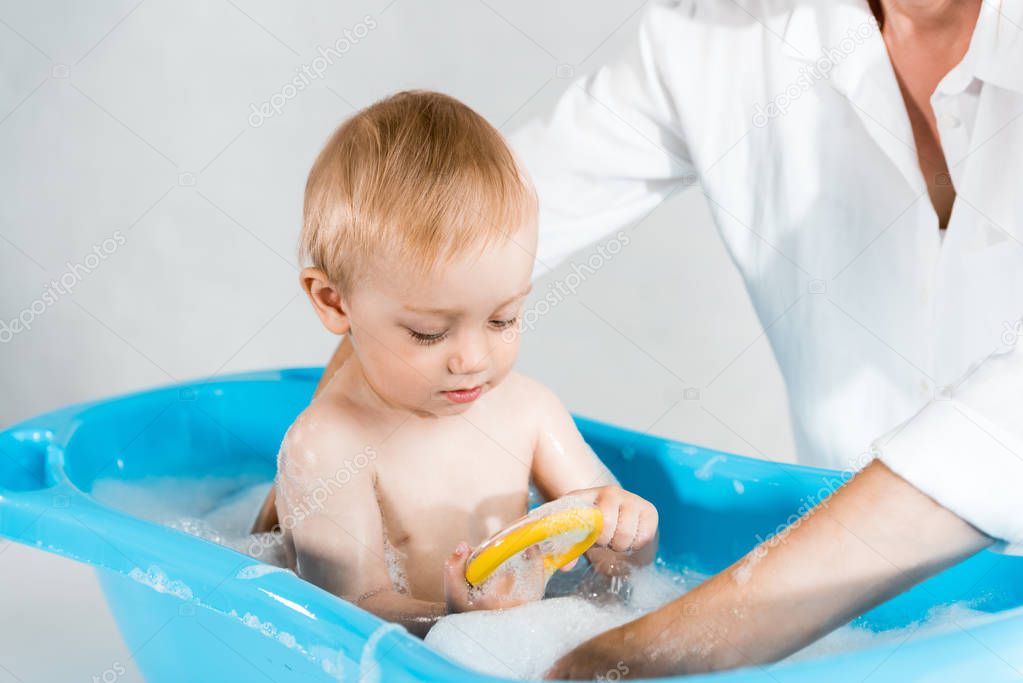 cropped view of mother washing adorable toddler kid in blue baby bathtub 