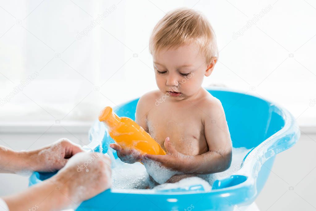 cropped view of mother near cute toddler child holding bottle with shampoo 