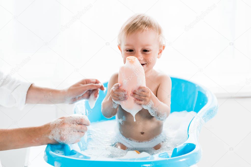 cropped view of mother near cheerful toddler child holding bottle with shampoo 