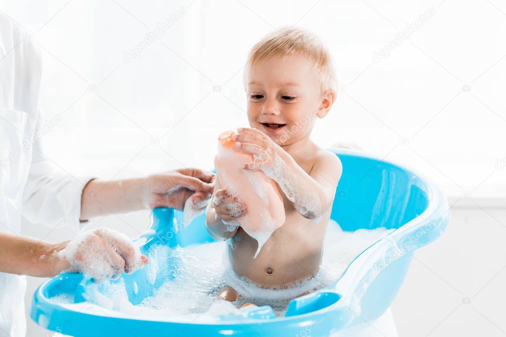 cropped view of mother near cheerful toddler son holding bottle with shampoo 