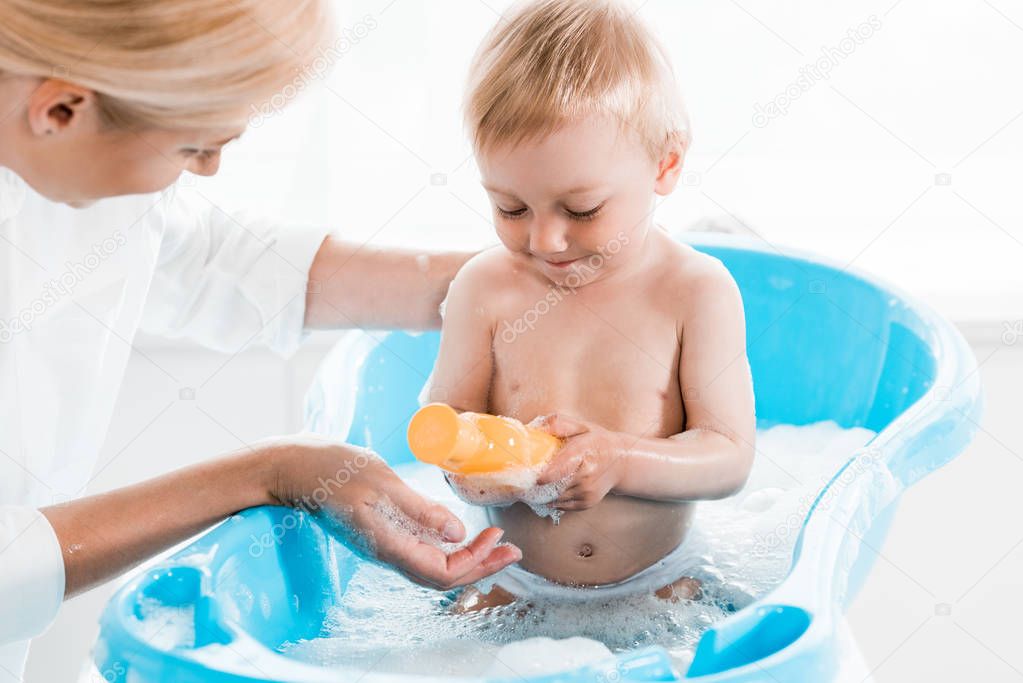 cropped view of blonde mother near cheerful toddler son holding bottle with shampoo 