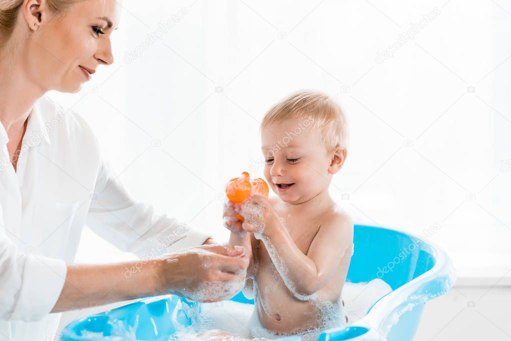 happy mother washing cheerful toddler son holding rubber duck in bathroom 