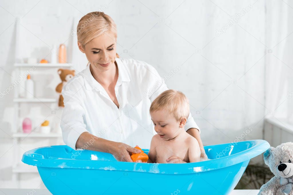 attractive blonde mother holding rubber toy near toddler son in plastic baby bathtub 