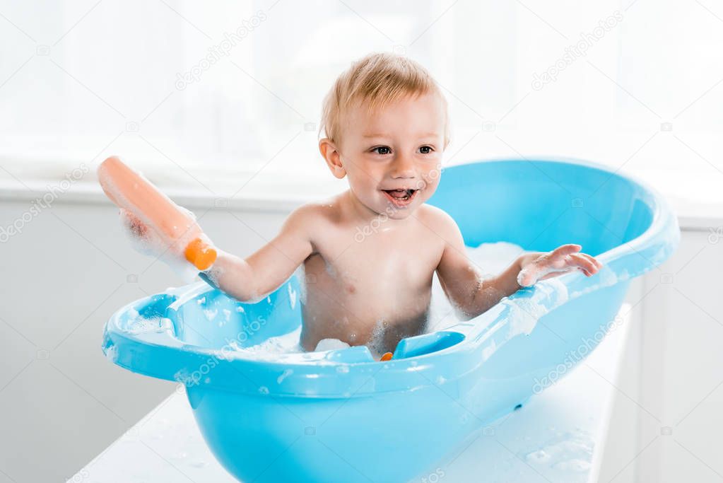 happy toddler kid smiling while taking bath in blue baby bathtub and holding bottle with shampoo 
