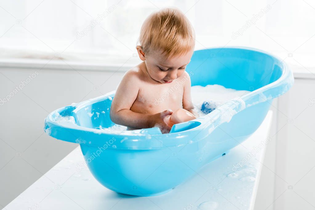 cute toddler kid looking at bottle with shampoo while taking bath in plastic baby bathtub 