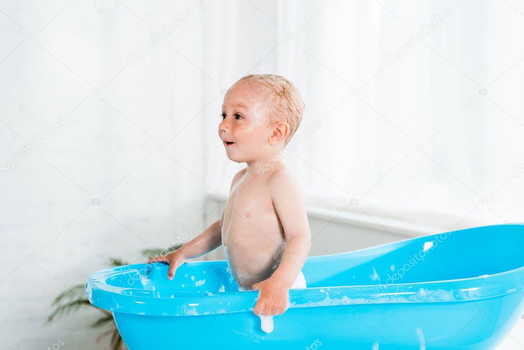 cute toddler kid in bath foam standing in plastic baby bathtub 