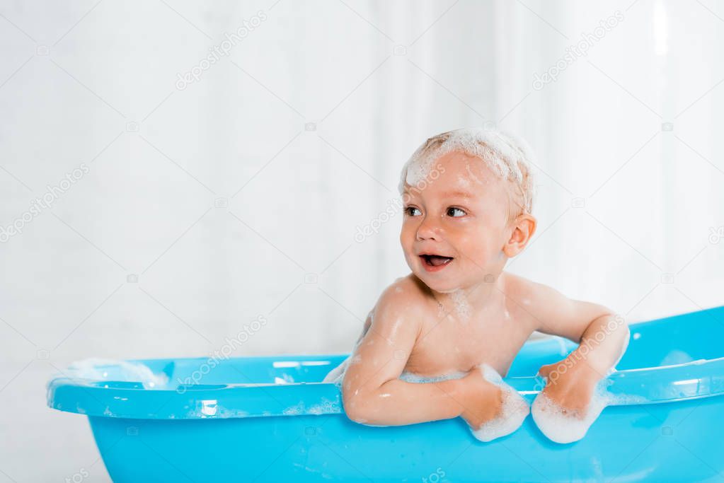 naked and cute toddler kid smiling while taking bath in plastic baby bathtub 